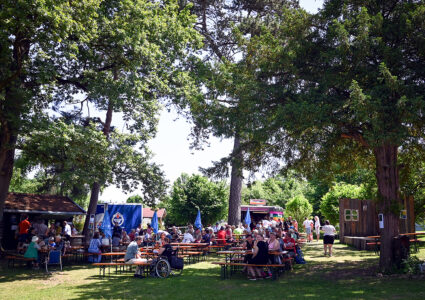 Feiern im Der Hermannsberg-Hotel - Zahlreiche Menschen sitzen auf Bierbänken zwischen den Bäumen. Im Hintergrund ist der Food-Trailer von Der Hermannsberg zu sehen.