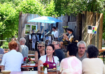 Feiern im Der Hermannsberg - Gäste verweilen glücklich auf im Schatten gelegenen Biergarnituren. Im Hintergrund spielt eine Blasmusikkapelle.