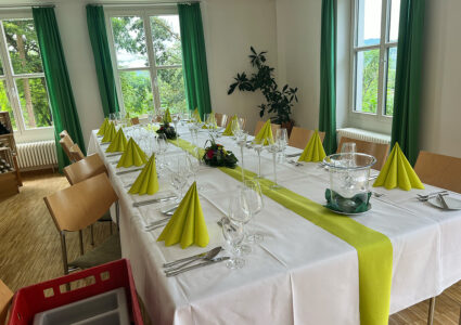Feiern im Der Hermannsberg - Gastraum mit festlich dekoriertem Tisch mit Gläsern, Tellern und Servietten. Durch die Fenster ist die idyllische Aussicht zu erkennen.